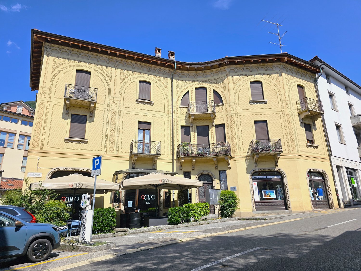 3-story building, yellow exterior, balconies on each floor, storefront with canopy, parking sign, greenery in front