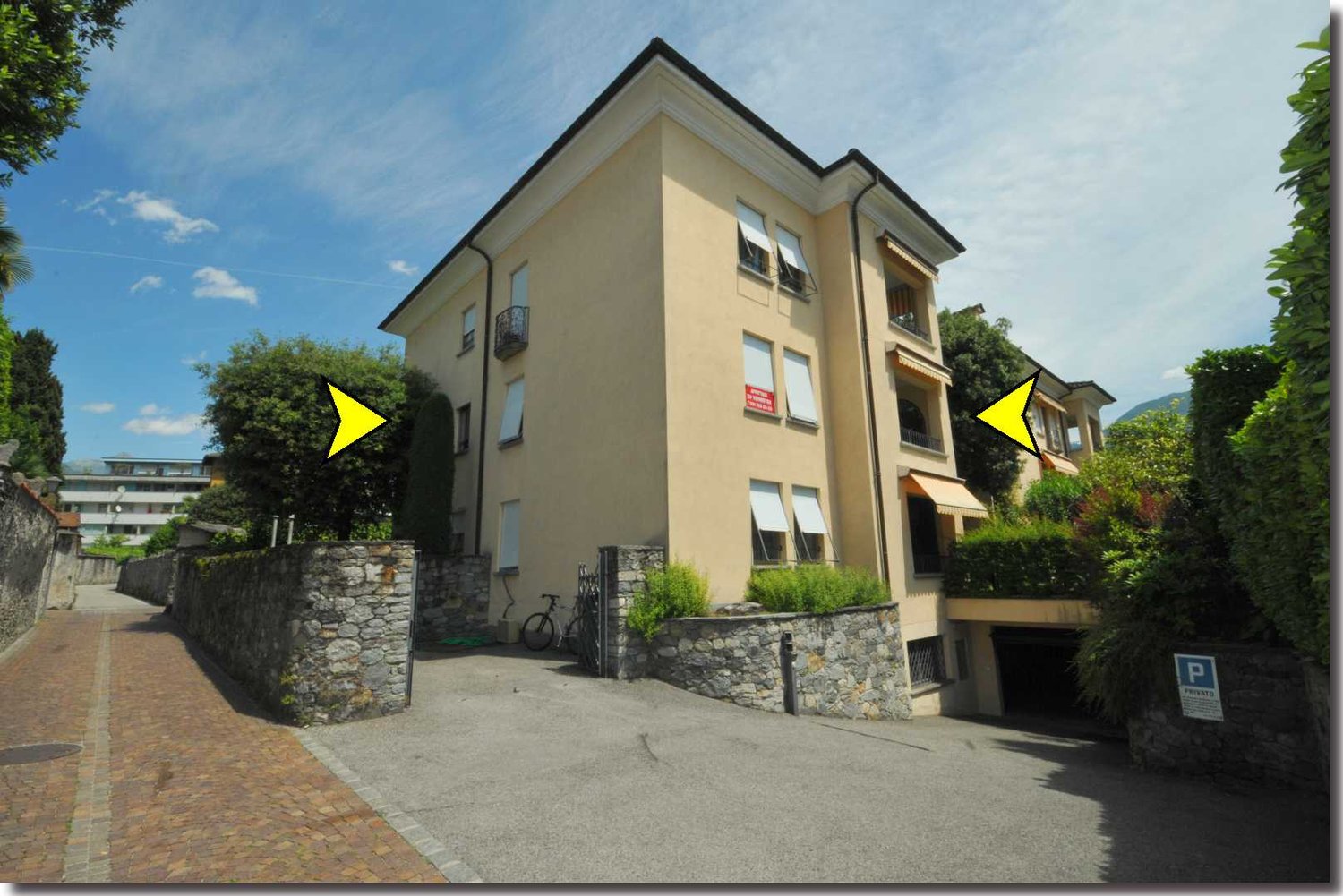 Yellow exterior, balconies with awnings, gated driveway, parking, bicycle, bushes, stone walls