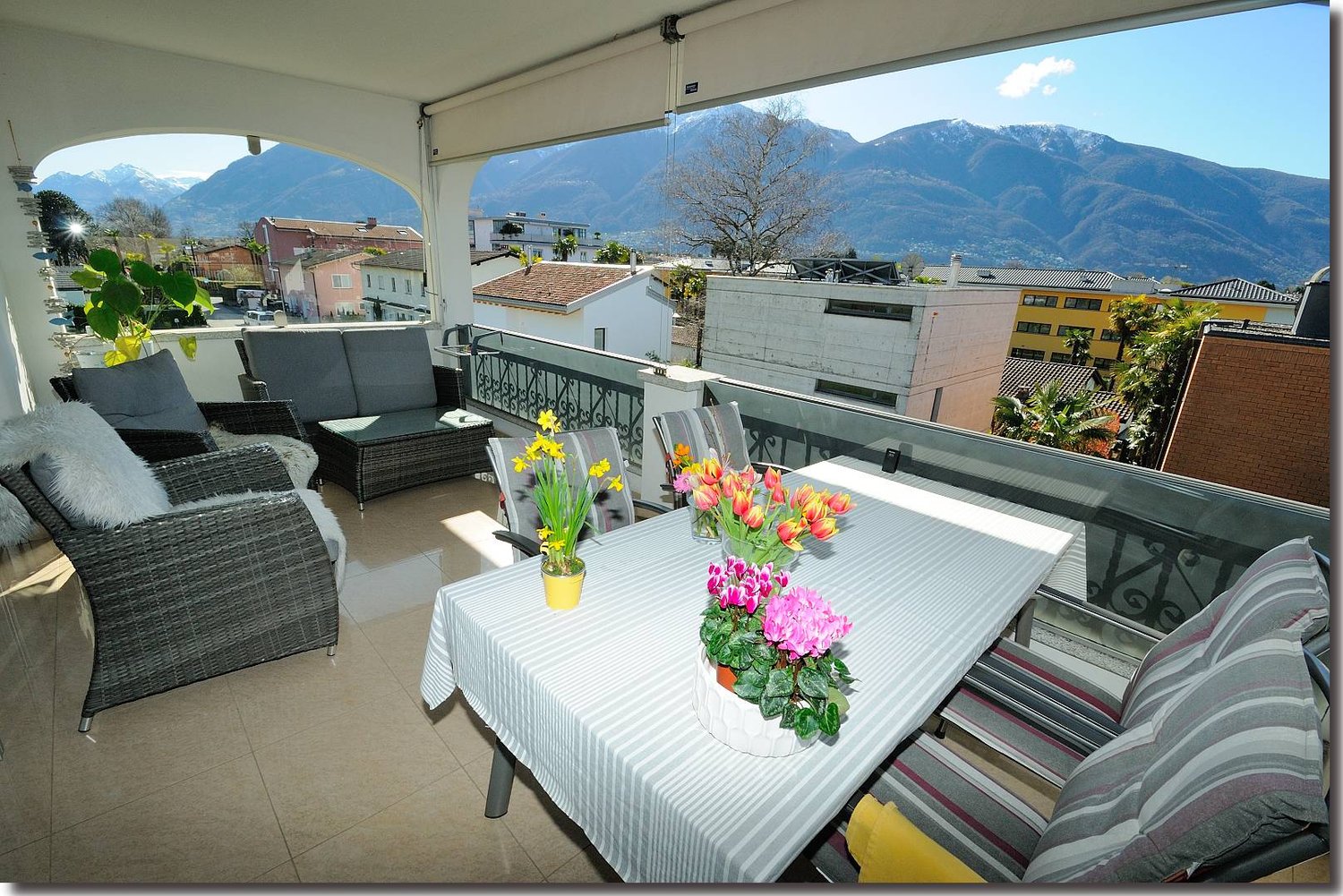Spacious balcony with a view of the mountains, furnished with wicker patio furniture, a dining table, and colorful flower arrangements. The balcony has a tiled floor and is surrounded by a railing.