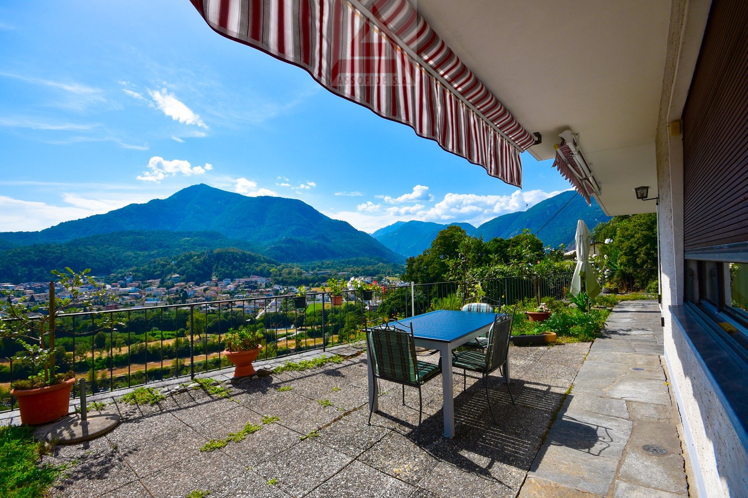 Balcony with a stunning view of the mountains and valley below. The balcony has a striped awning, a table and chairs, and potted plants. The surrounding landscape is lush and green, with mountains in the distance under a blue sky with clouds.