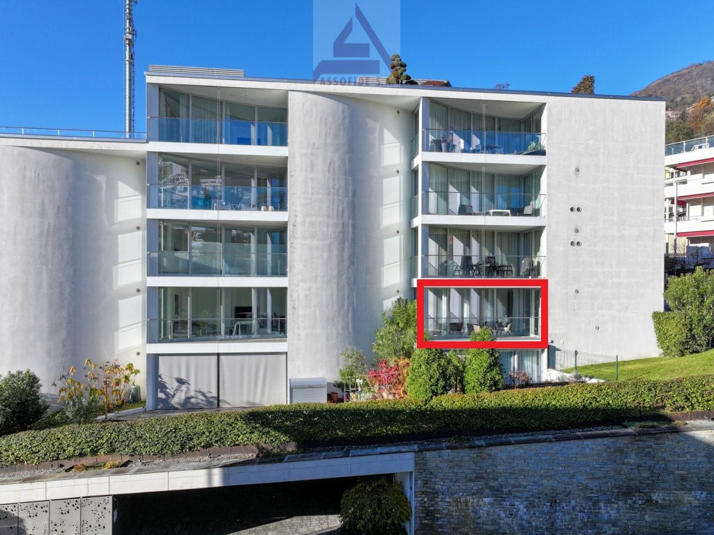 Modern multi-story apartment building with glass balconies and terraces, surrounded by landscaping and a water feature.