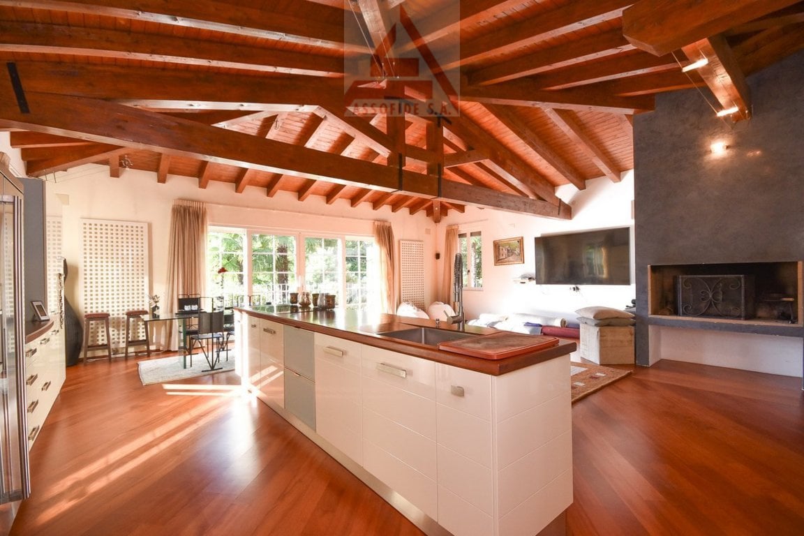 Large open-concept living space with wooden beamed ceiling, hardwood floors, kitchen with white cabinets and wooden countertops, living area with fireplace, and large windows overlooking the outdoors.