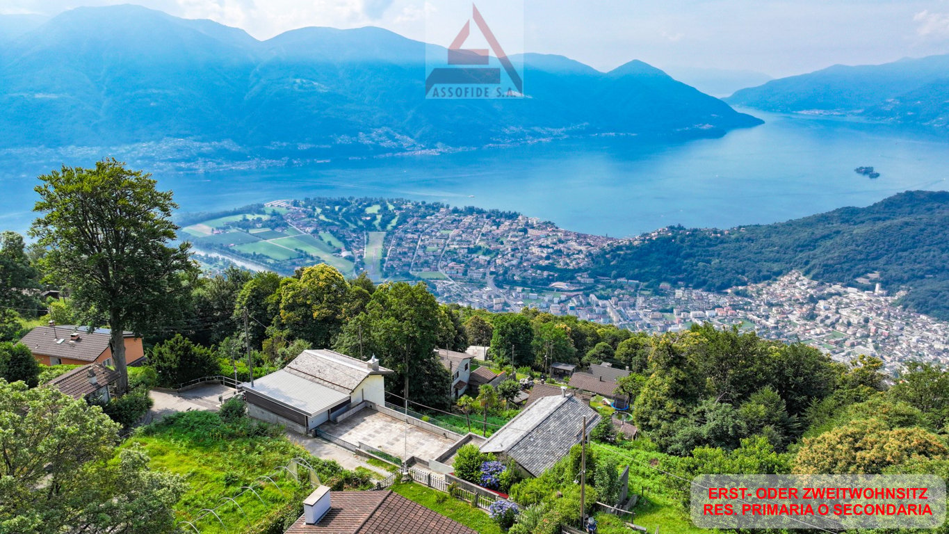 Aerial view of a mountainous town with a lake in the background. The town is surrounded by lush green trees and vegetation. There is a large sign for 'Assofide S.A.' visible in the image.