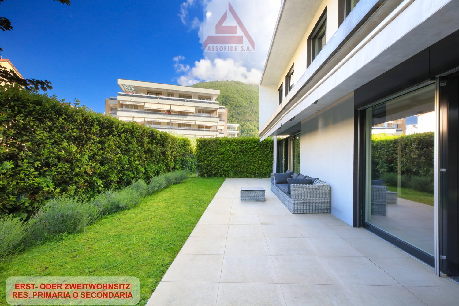 A white building with glass windows, tiled patio leading to a sofa on a terrace, green hedges and grass lawn, and a mountain in the background.