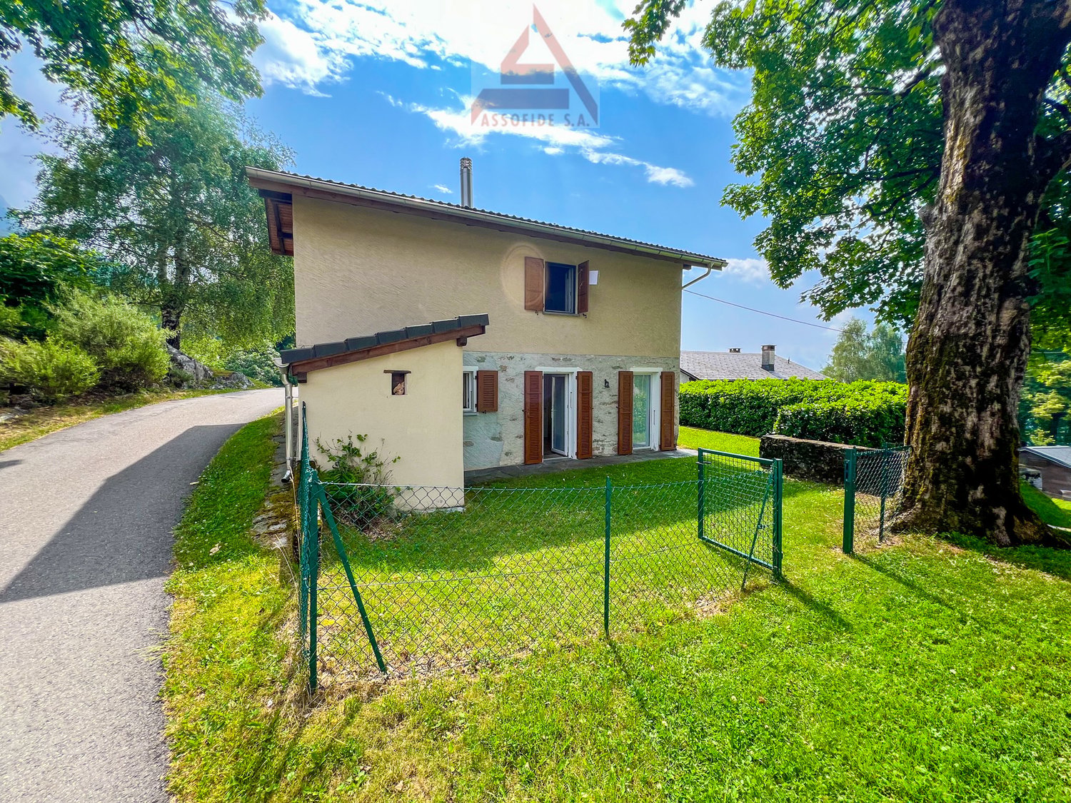 Detached house, beige exterior, green fence, driveway, garden, trees, bushes, clear sky