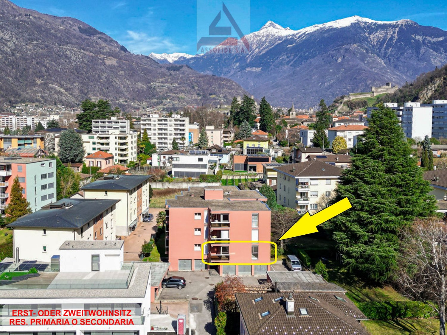 Aerial view of a residential area with a highlighted apartment building, surrounded by other buildings, trees, and mountains in the background.
