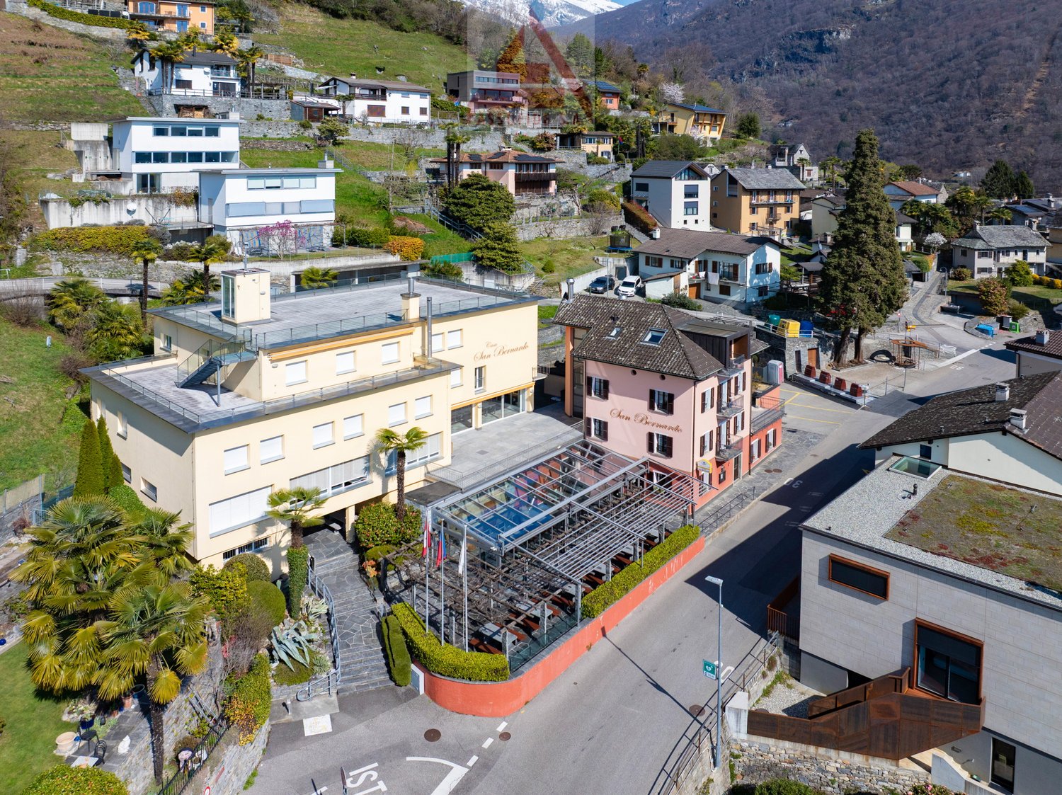 Aerial view of a hillside village with multiple buildings, some with glass roofs, surrounded by greenery and mountains in the background.