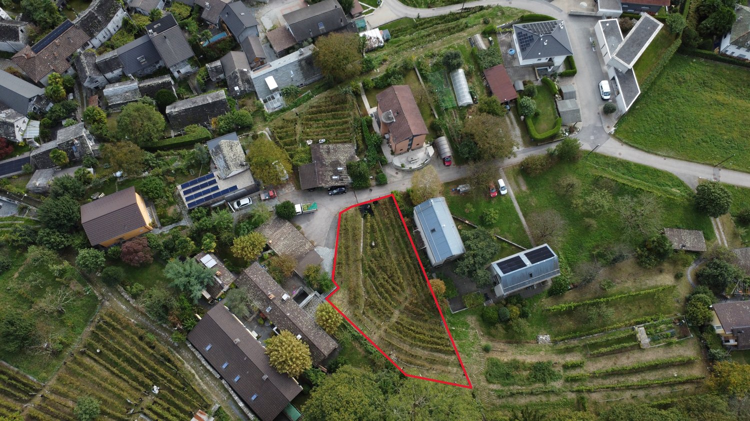 This image shows an aerial view of a residential area with a mix of houses, buildings, and gardens. There are several small plots of land or gardens visible, some of which appear to be used for growing crops or vegetables. There is also a parking area or