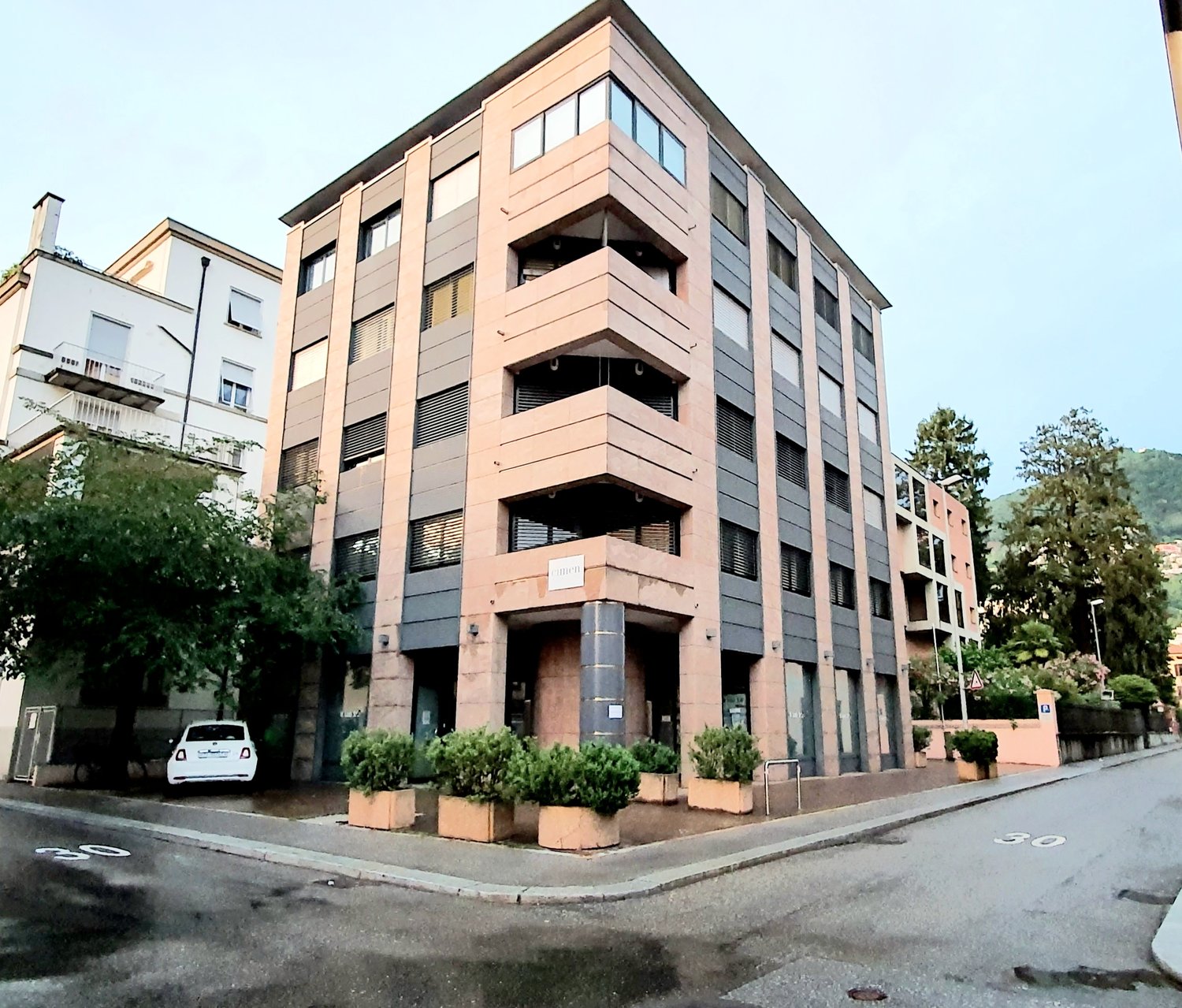Multi-story building, beige and gray exterior, white car parked in front, many windows, plants outside