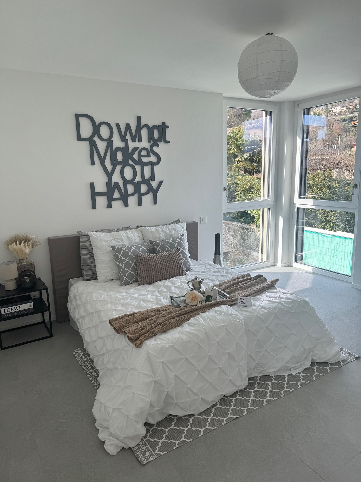 large bed with white duvet, grey and white pillows, black nightstand with books, lamp, round white lampshade, large windows overlooking pool