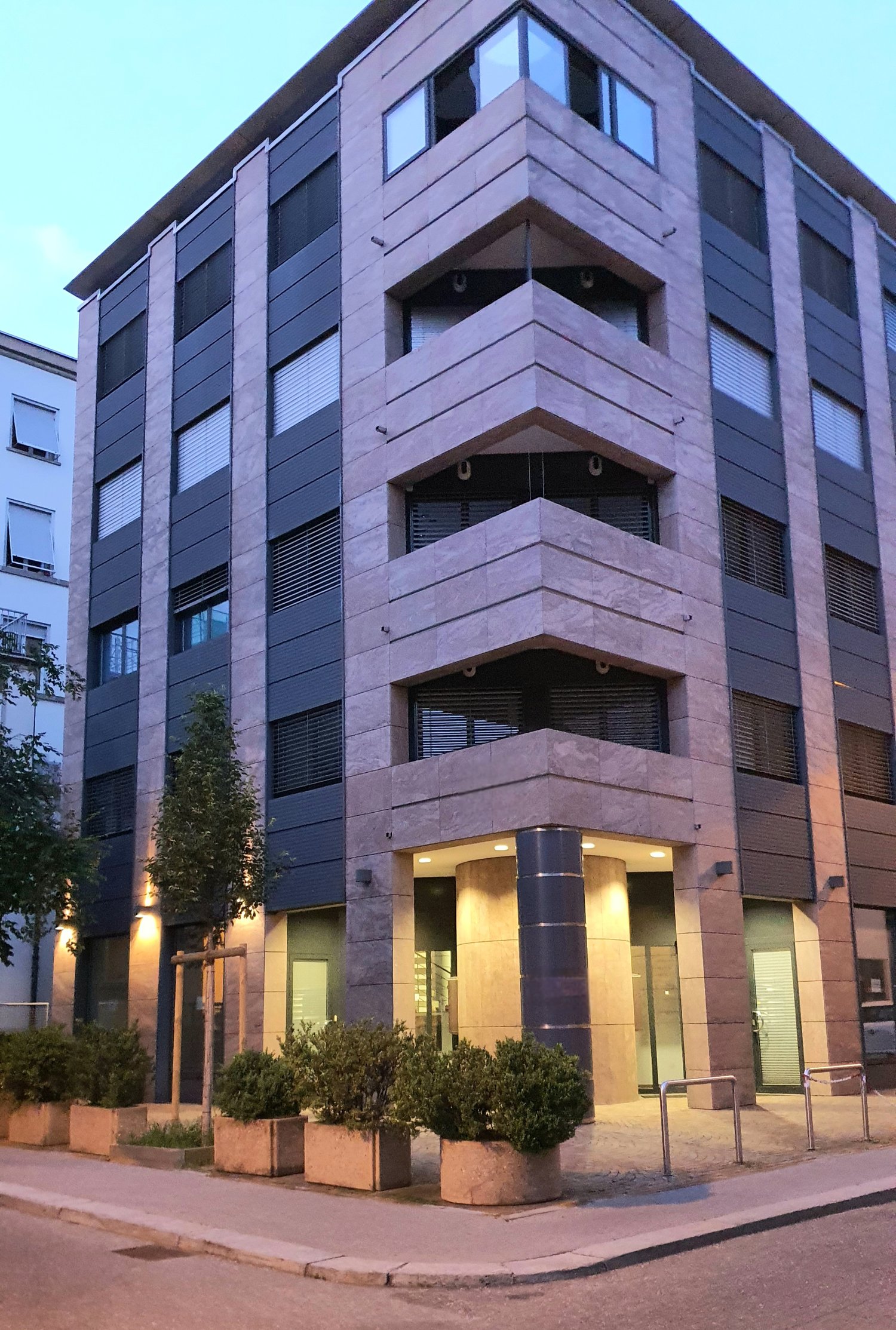 Modern apartment building, multiple floors, glass windows, entrance with lights, shrubs and trees in front