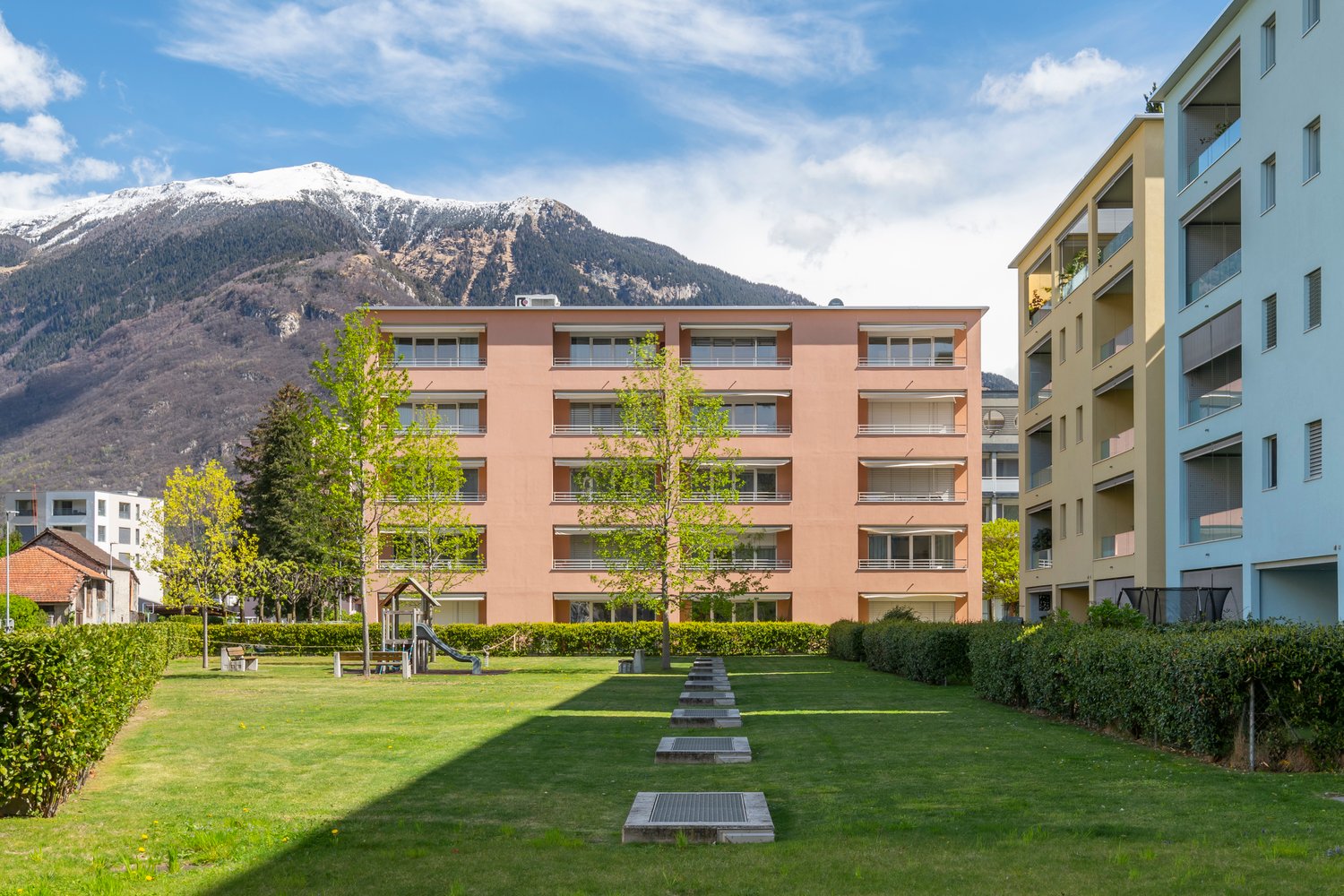Two story apartment building, white and pink exterior, several balconies, green lawn, bushes, playground, trees, mountains in the background