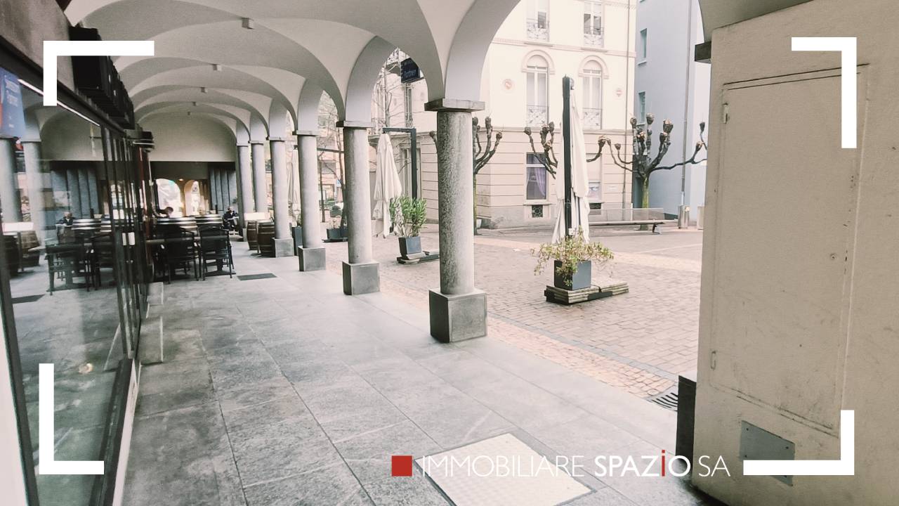 The image shows an outdoor area with arched walkways, columns, and potted plants. The ground is paved with stone tiles, and the surrounding buildings have a historic architectural style. This appears to be a commercial or public space, potentially a plaza