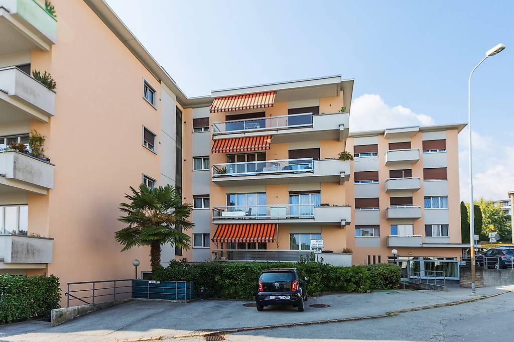 Multi-story apartment building with balconies, red and white exterior, palm tree, parking space in front