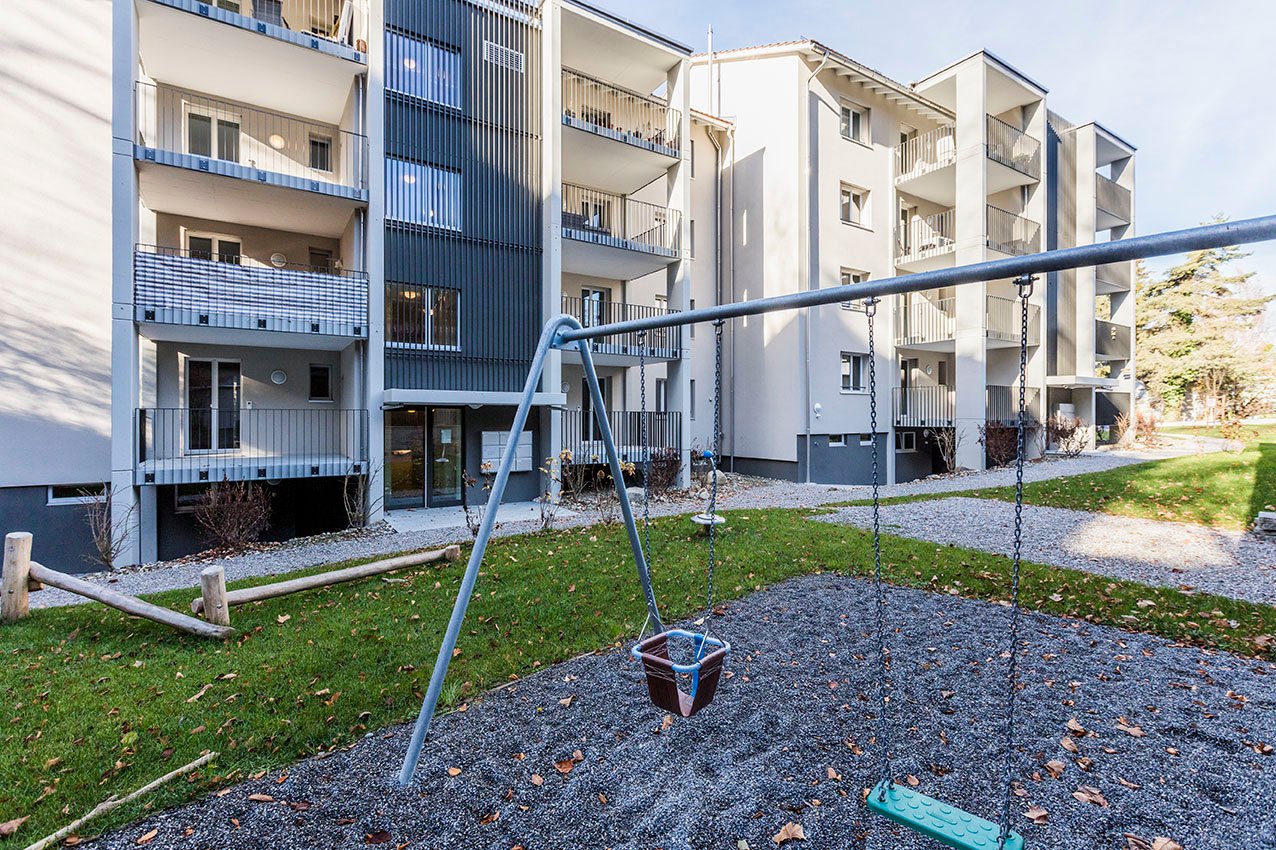 Modern apartment building, three floors, balcony, metal railings, entry way, swing set, gravel area, grassy area, trees