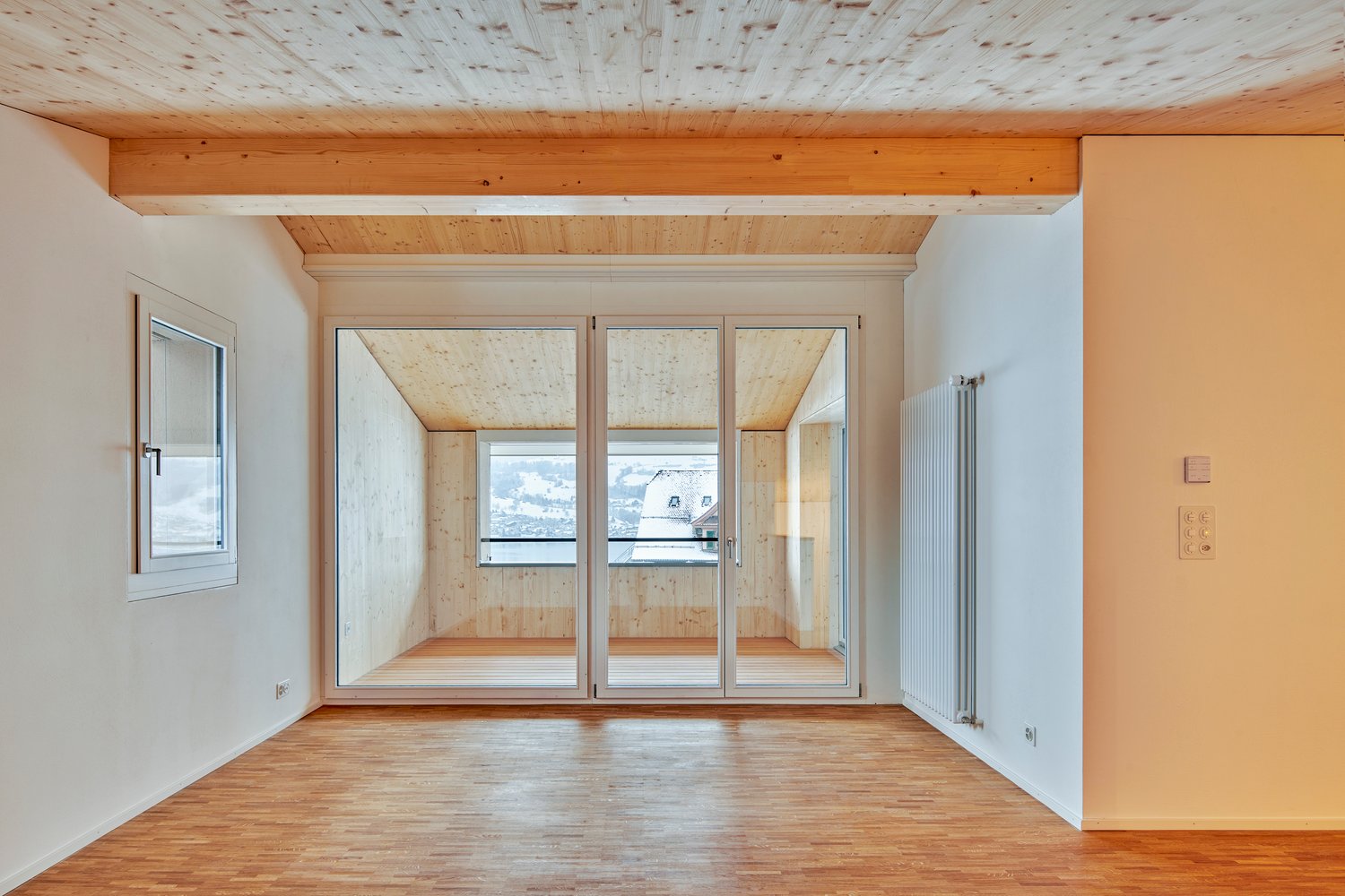 Empty room with hardwood floors, large sliding doors leading to a balcony, exposed wooden ceiling, small window on the left, radiator on the right wall.