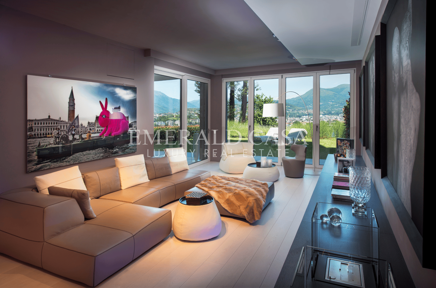 Spacious living room with large windows offering a scenic view of the mountains, modern furniture including a beige sectional sofa, a glass coffee table, and decorative accents such as a pink rabbit sculpture and a wall art piece depicting a cityscape.