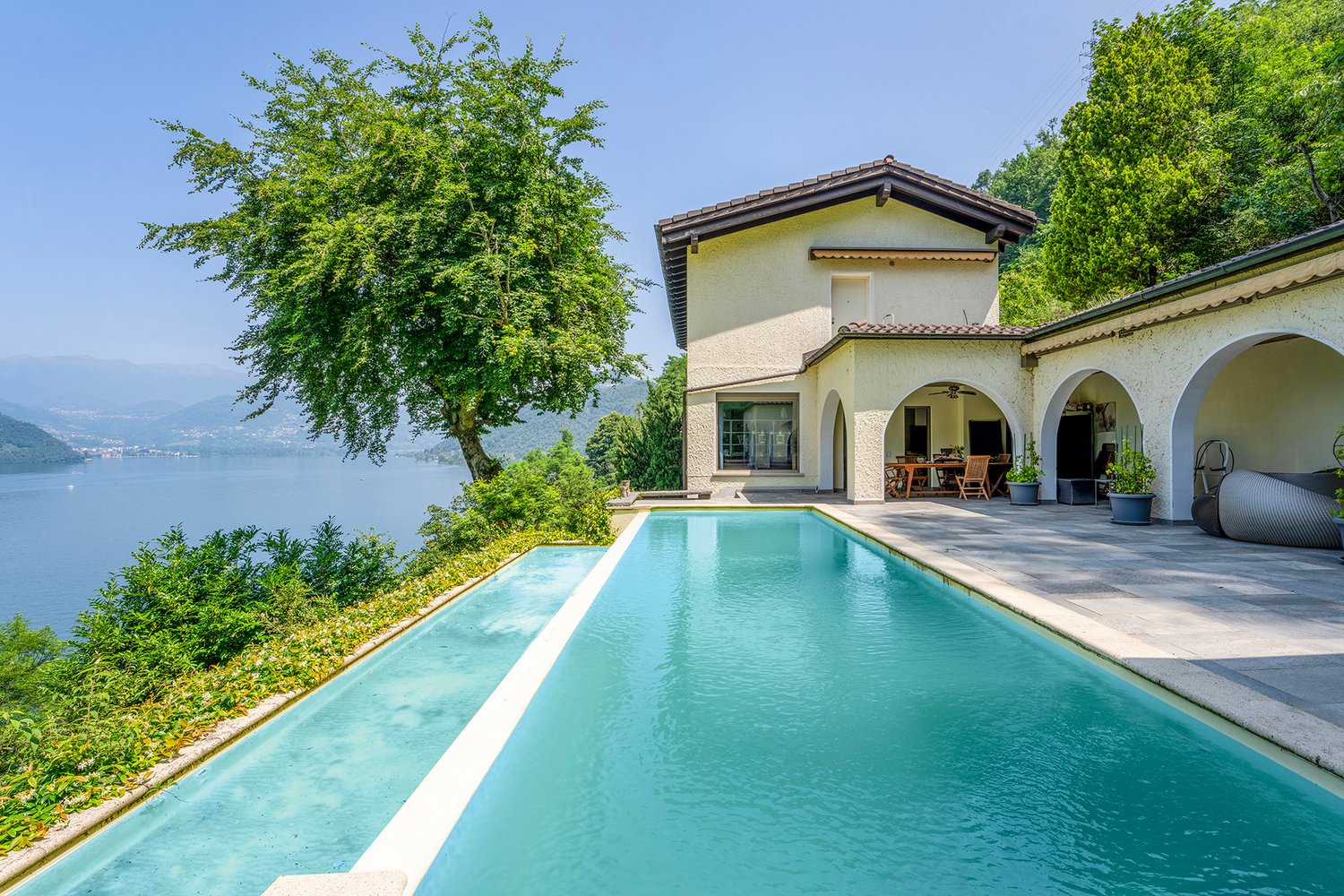 Two-story villa with a swimming pool, surrounded by lush greenery and overlooking a lake. The villa has a terrace and large windows, providing a scenic view of the natural landscape.