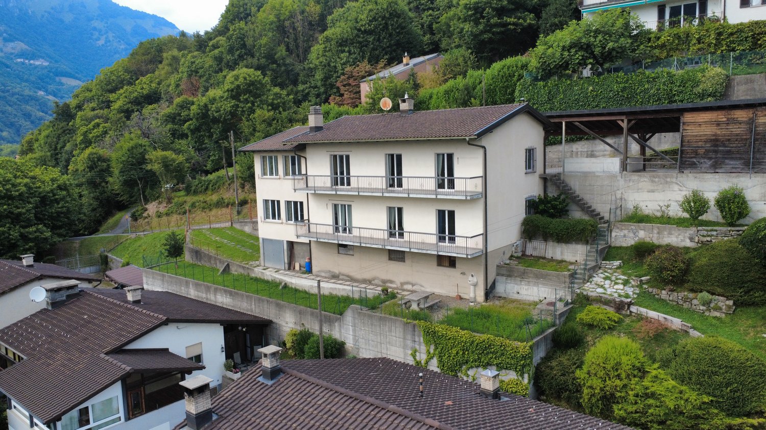 Detached house, multiple floors, tiled roof, exterior balconies, walled terrace, neighboring houses visible, situated on a slope with retaining walls