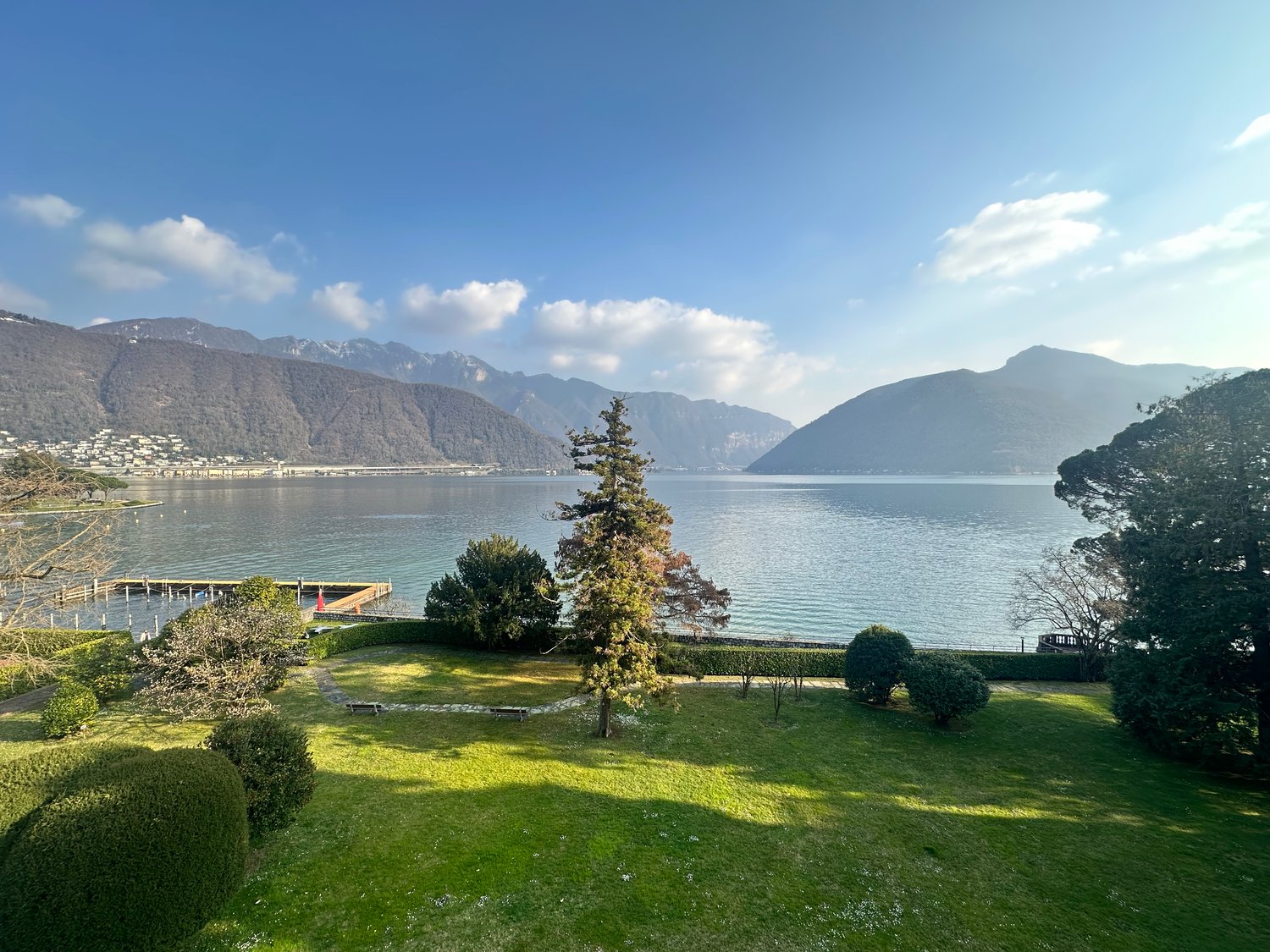 Green lawn, various trees and bushes, distant mountains, lake with a dock