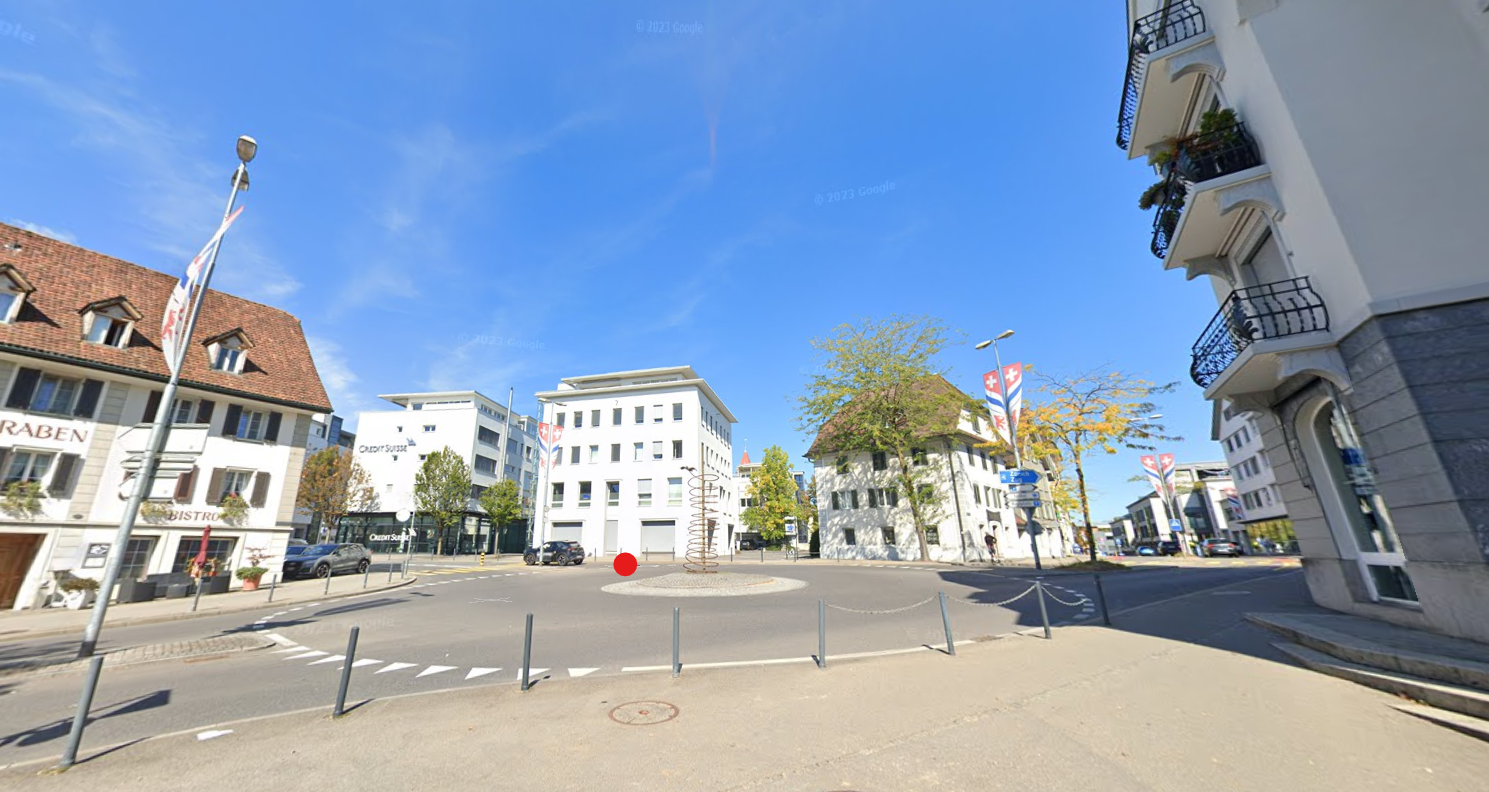 The image shows a street view of a city center. There are various buildings, including office buildings and shops, lining the street. The street has a roundabout in the center, and there are parked cars and pedestrians visible. The buildings have a mix of