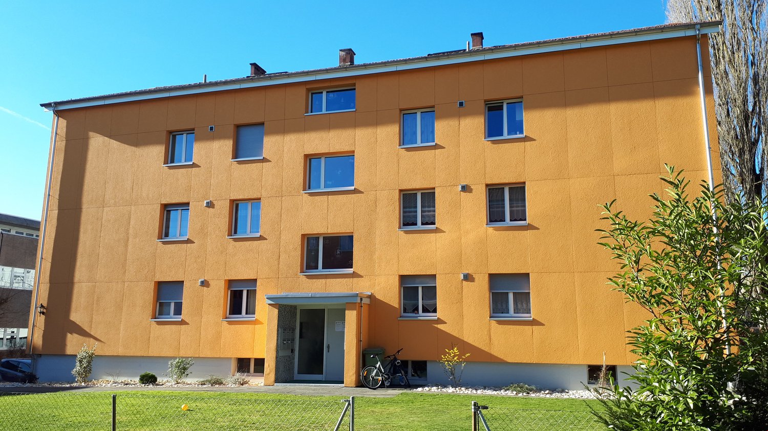 4 story apartment building, yellow walls, many windows, entrance at the bottom, bicycle parked outside, fence, shrubs