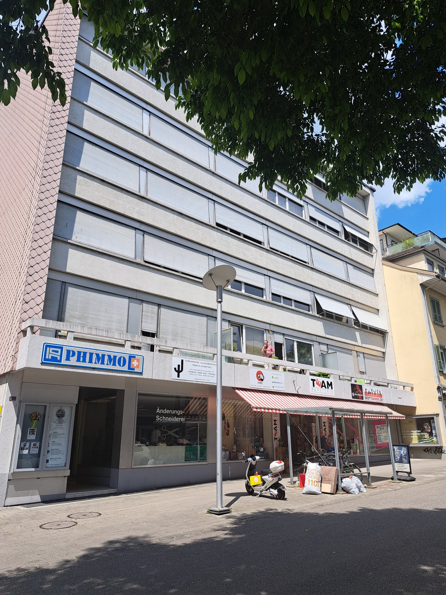 A building with several floors, exterior paint in pink and white, entrance with door and window, ground floor shops with signs, awnings, and signage, street light and parked motorbike