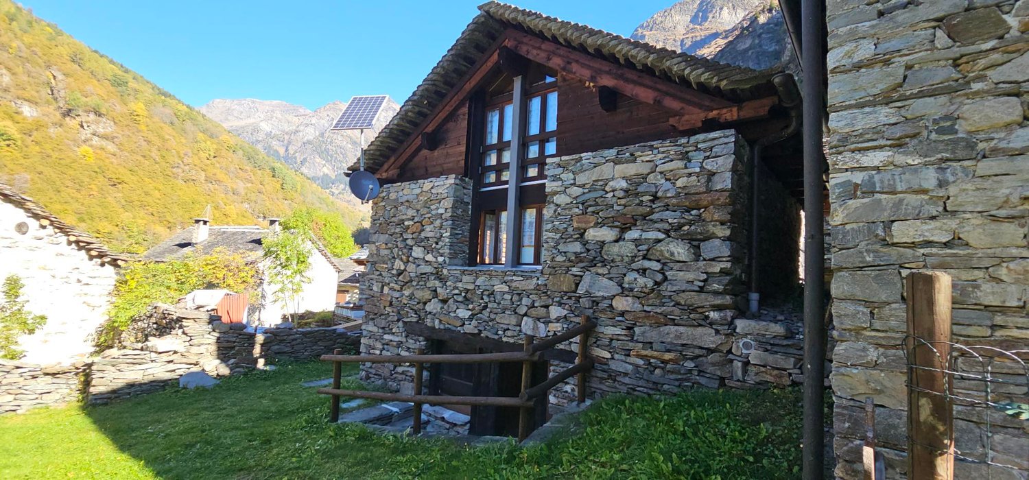 Old house made of stone, with a solar panel on the roof, wooden door, two windows, surrounded by lush green grass