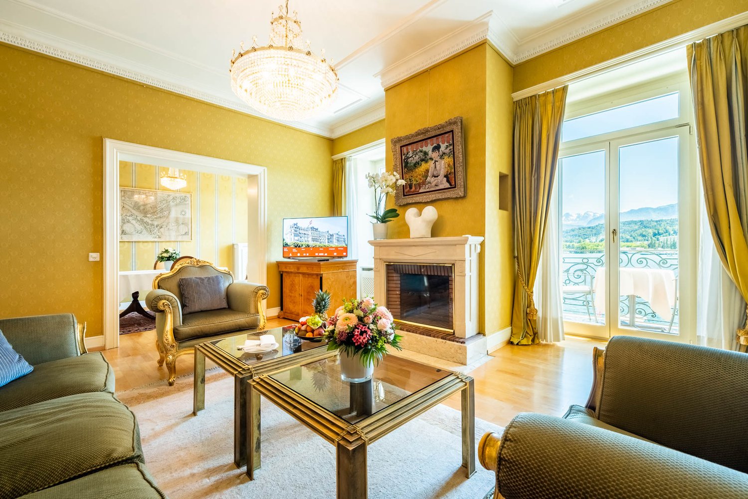 Spacious living room with yellow walls, ornate chandelier, large windows with balcony access, fireplace, plush seating, and decorative artwork and floral arrangements