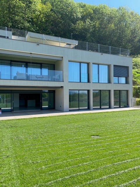 Modern multi-story building with large glass windows, balconies, and a grassy lawn in the foreground.