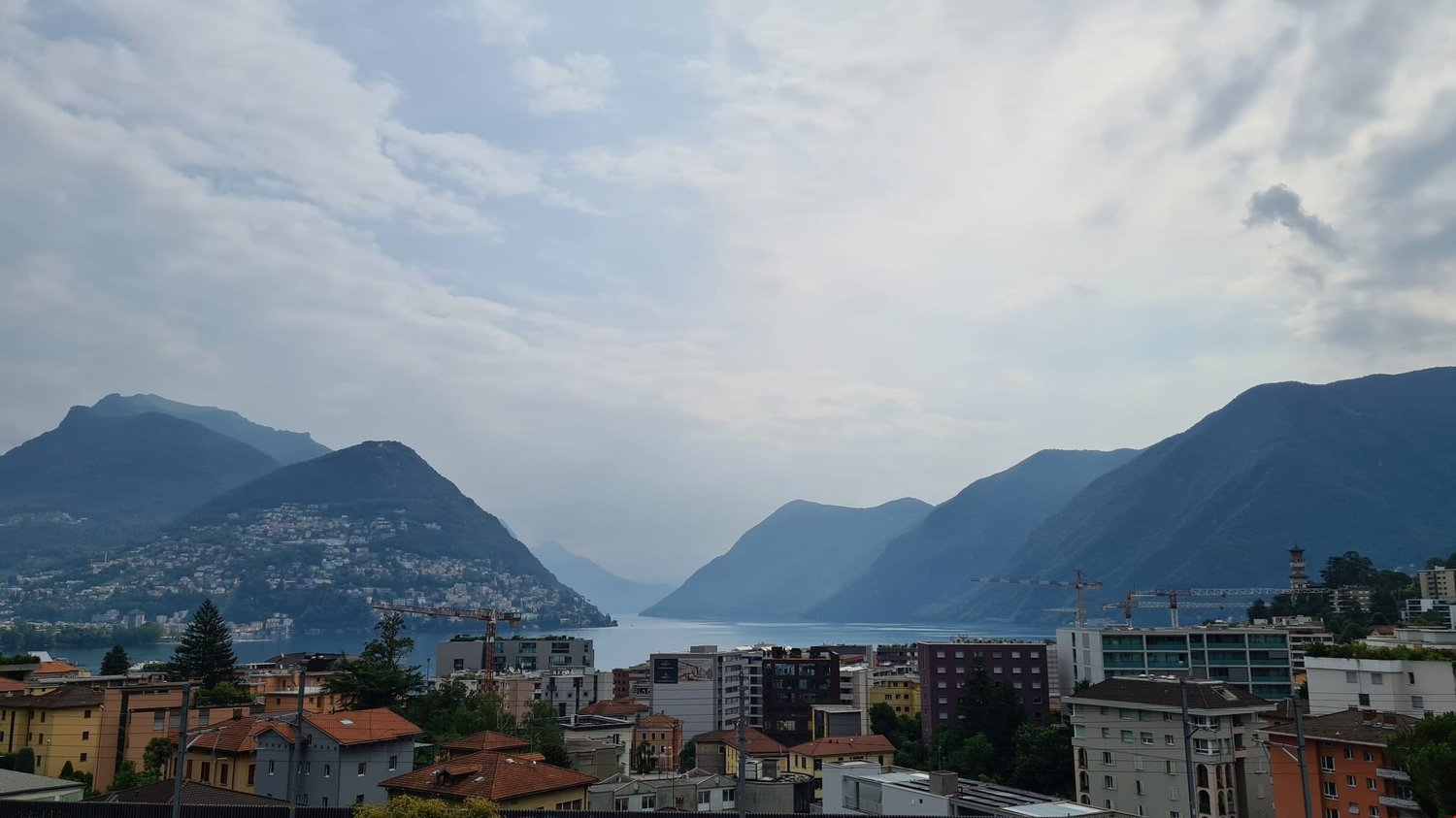 Wide panoramic view of mountainous region, water body, urban buildings, cranes, trees