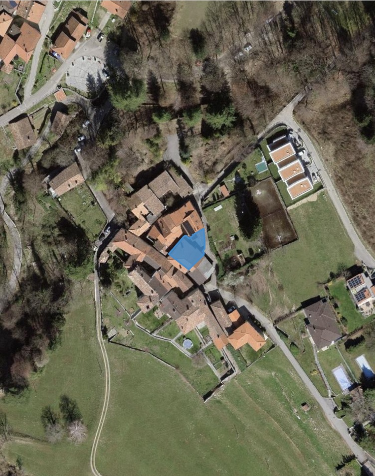 The building is an old medieval-style stone house located in the village. The surrounding area is rural, with houses, farmland, and some trees. A blue arrow points to the main entrance of the building.