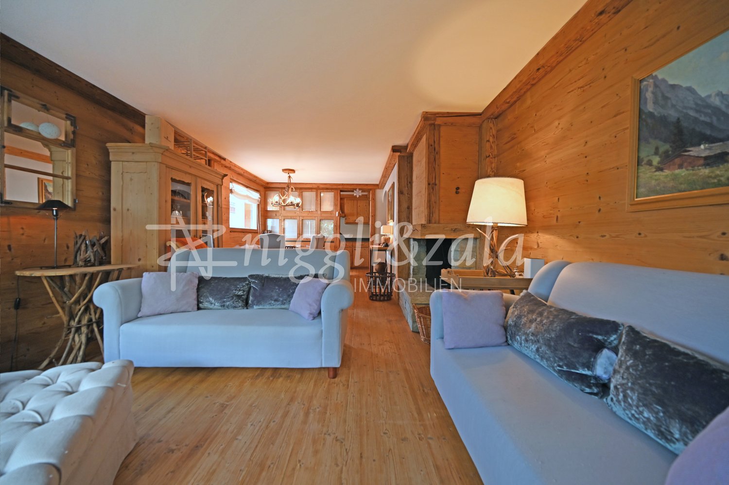 2 sofas, 1 armchair, wooden floor, wooden wall, fireplace, ceiling light, lamps, wooden table, wooden cupboard, paintings, chandelier