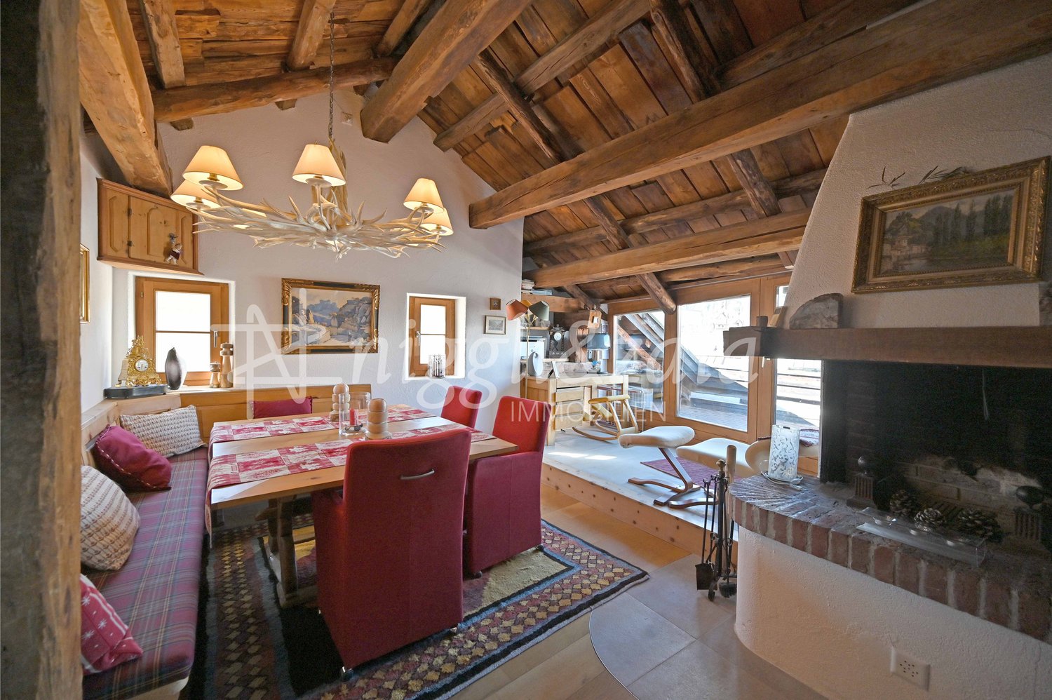 Dining area with dining table, red chairs, fireplace, wood beam ceiling, large windows, wood rocking chair, chandelier
