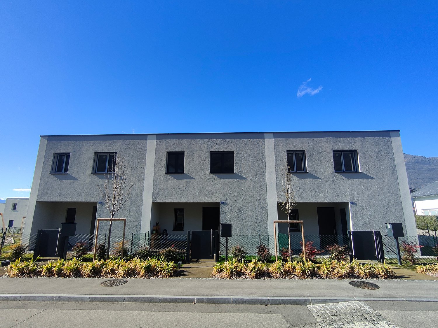 Modern two-story residential building with grey walls and black windows, gated front entrance, and shrubs in front