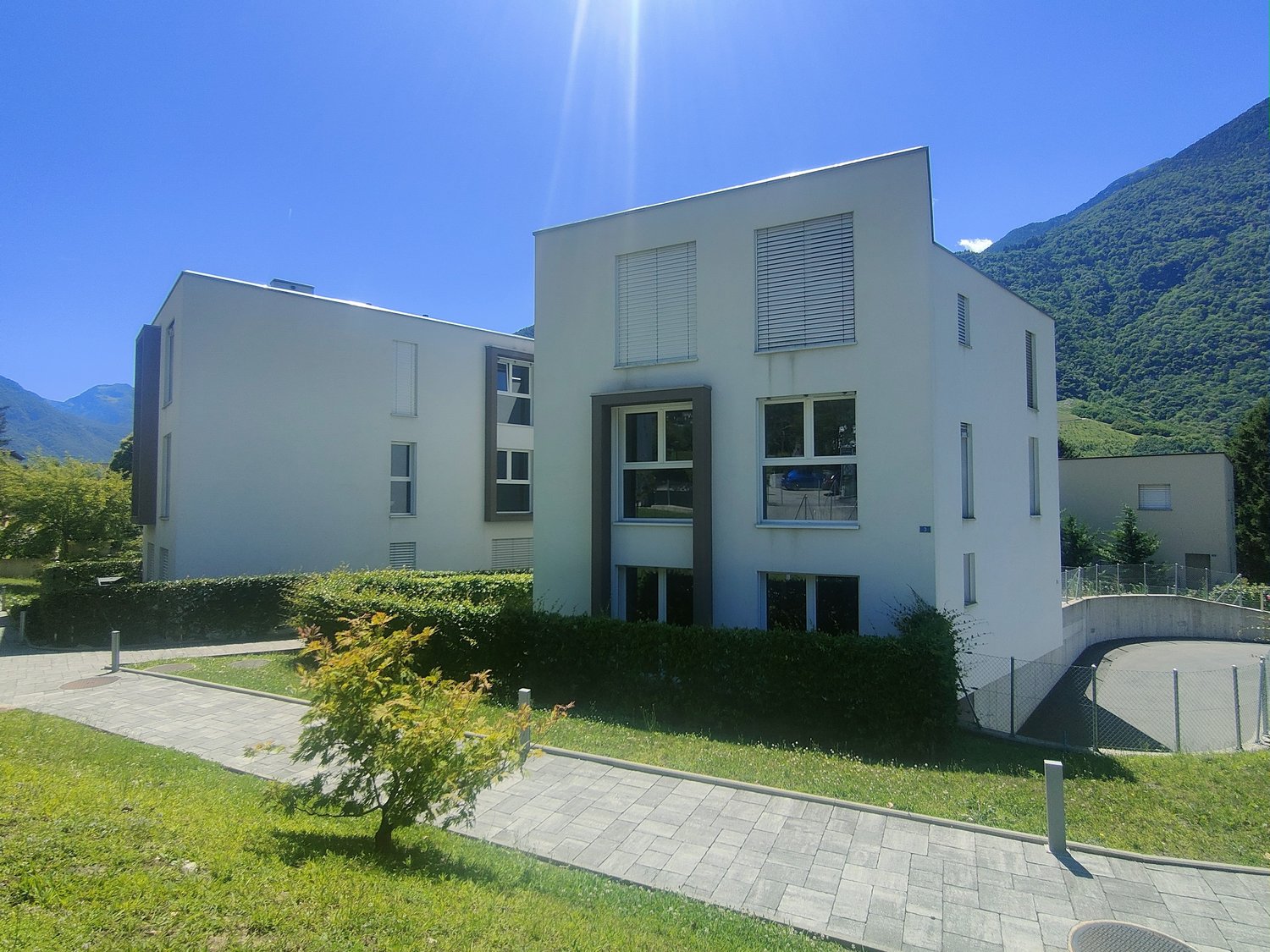 A modern, multi-story building with a minimalist, rectangular design. The building has large windows, a paved walkway, and a grassy area surrounding it. The building appears to be situated in a mountainous, forested area with a clear blue sky overhead.
