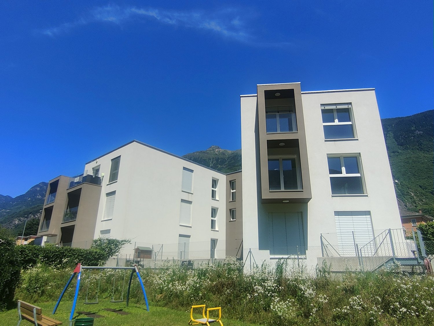 Multi-story apartment building with modern, minimalist design featuring large windows, balconies, and a grassy area with playground equipment in the foreground. The building is set against a backdrop of mountains under a clear blue sky.