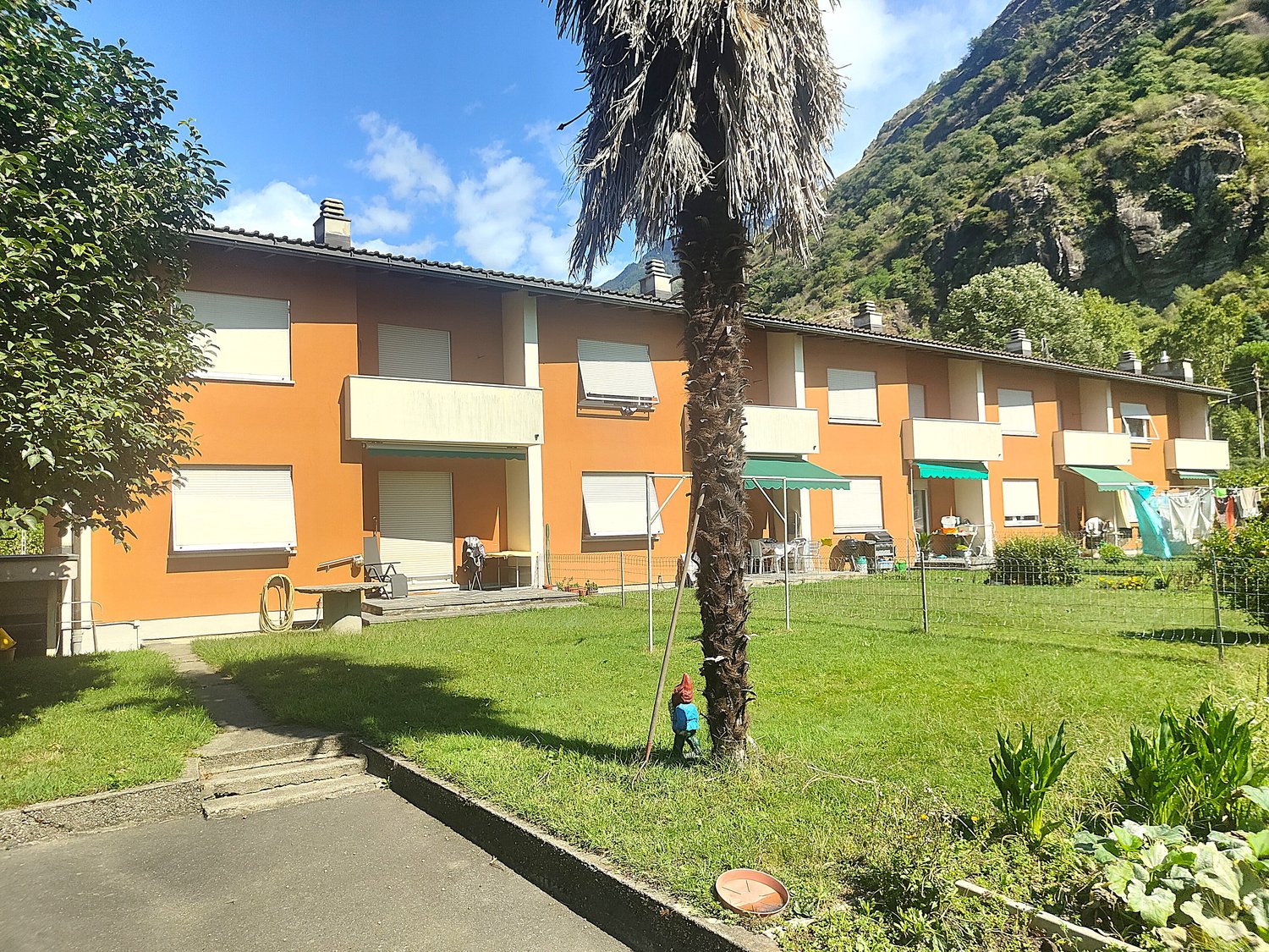 A multi-unit residential building with orange and white exterior, surrounded by a grassy lawn and palm trees. The building has balconies or terraces on each unit.