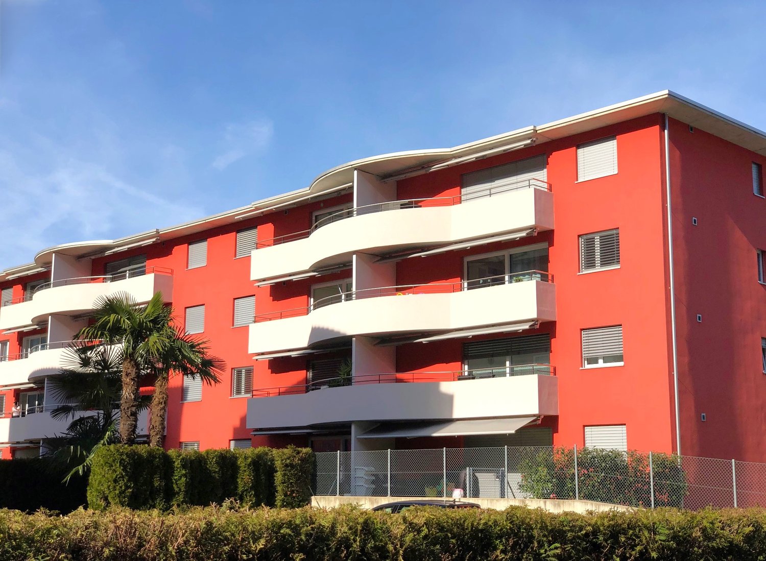 Multi-story apartment building with red and white exterior, balconies, and palm trees in the foreground