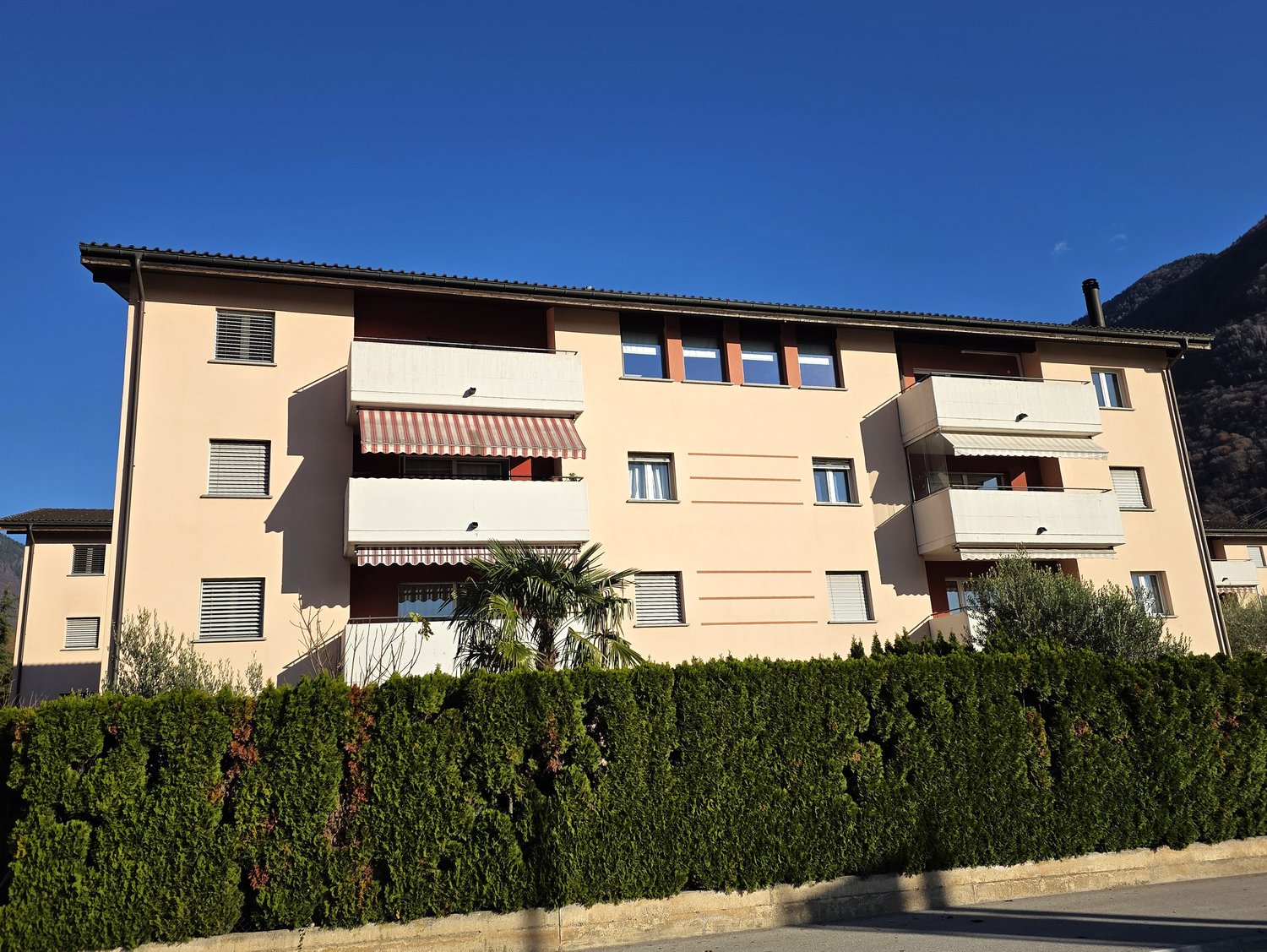 3 story apartment building with balconies and awnings, beige exterior with vertical stripes, green shrubs and palm trees
