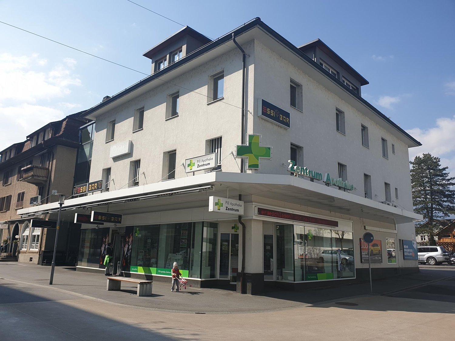 A multi-story commercial building with a pharmacy and medical center on the ground floor. The building has a gray exterior with large windows and a green cross symbol indicating the medical center. There is a person walking in front of the building.