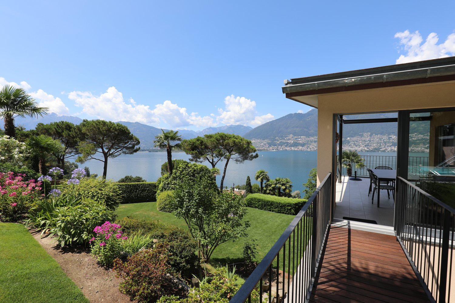 Luxury villa with a panoramic view of a lake surrounded by mountains. The property has a large balcony or terrace overlooking a well-manicured garden with palm trees, shrubs, and colorful flowers. The villa appears to be situated on a hillside, providing
