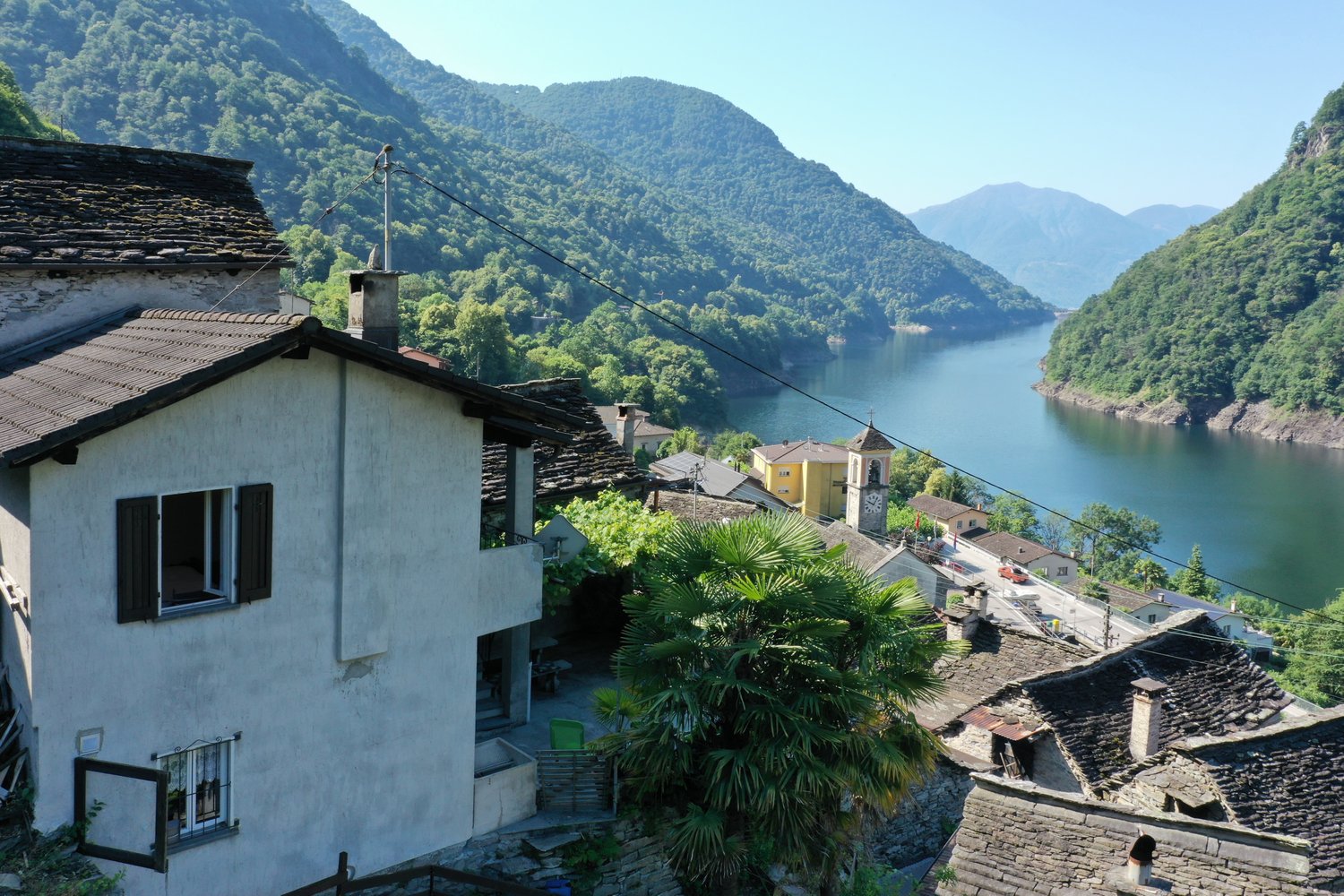 Picturesque village nestled in the mountains, with traditional stone and stucco buildings, palm trees, and a scenic lake in the background.