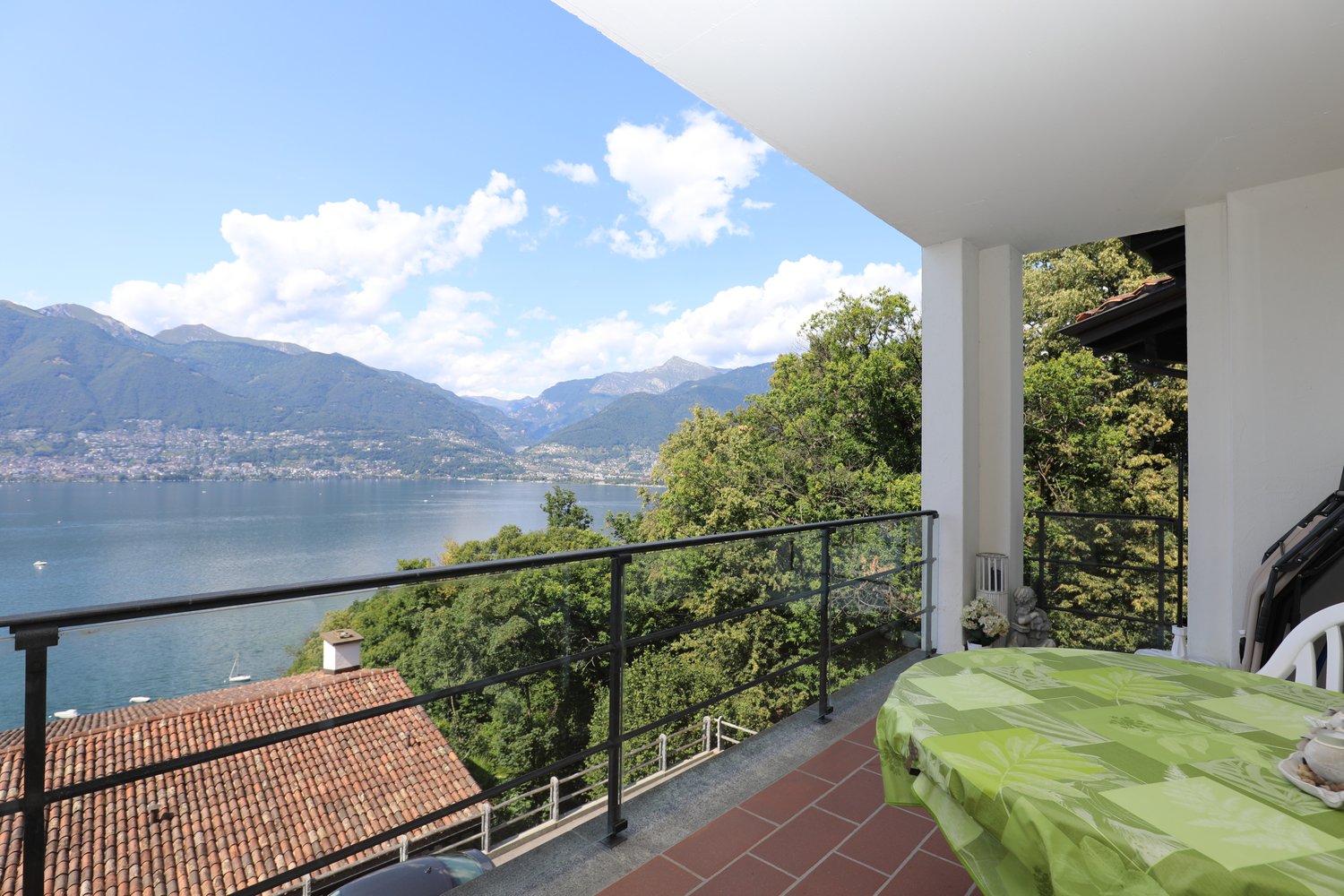 Spacious balcony with a stunning view of the lake and mountains. The balcony has a tiled floor and a metal railing. There is a table with a green tablecloth on the balcony, suggesting it is used for outdoor dining or relaxation.