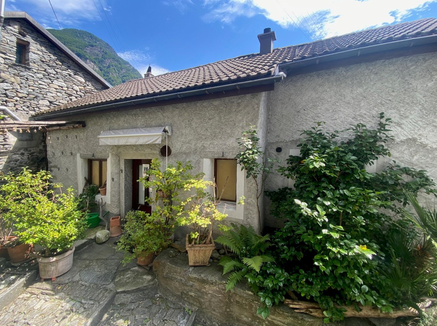 Stone house, brown roof, white windows, plants, pathway, mountain in the background