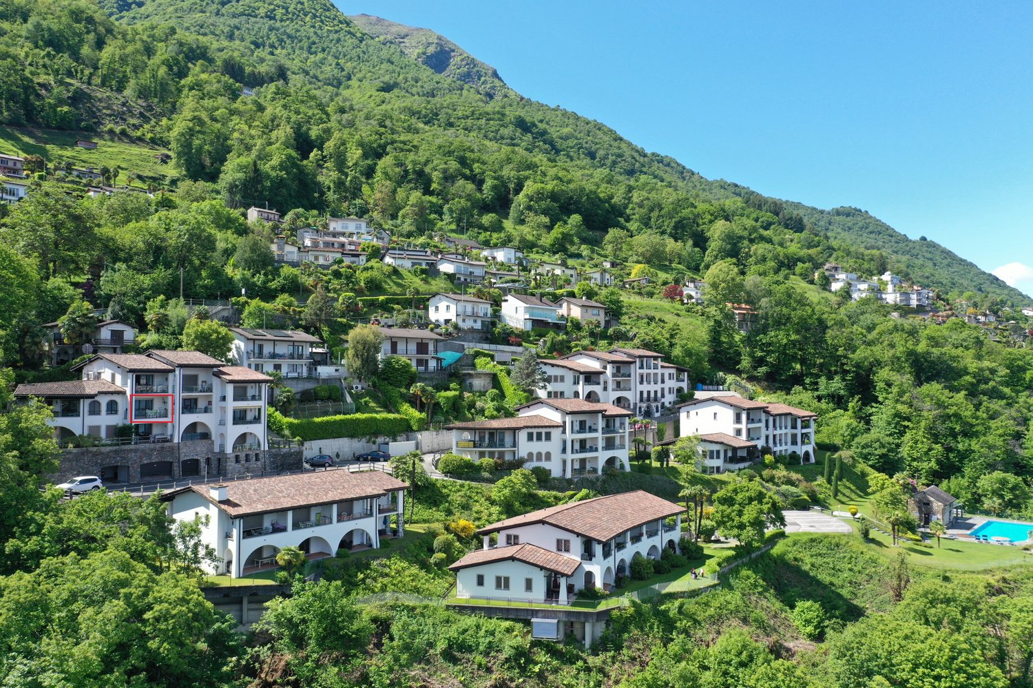 Hillside residential area with multi-story buildings, balconies, and parking spaces surrounded by lush green vegetation and mountains