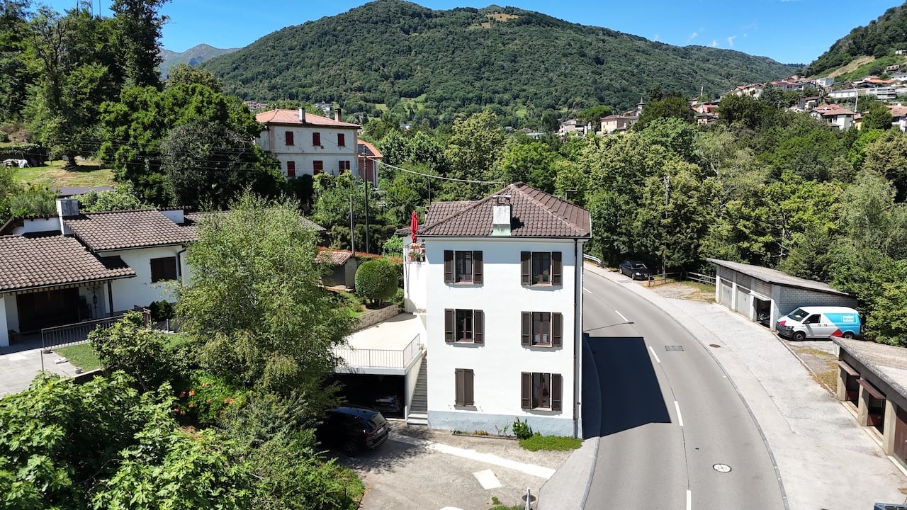 The image shows a small village nestled in a mountainous, forested area. There are several multi-story residential buildings with red tile roofs, surrounded by lush greenery. A road runs through the village, with a parked vehicle visible. The landscape is