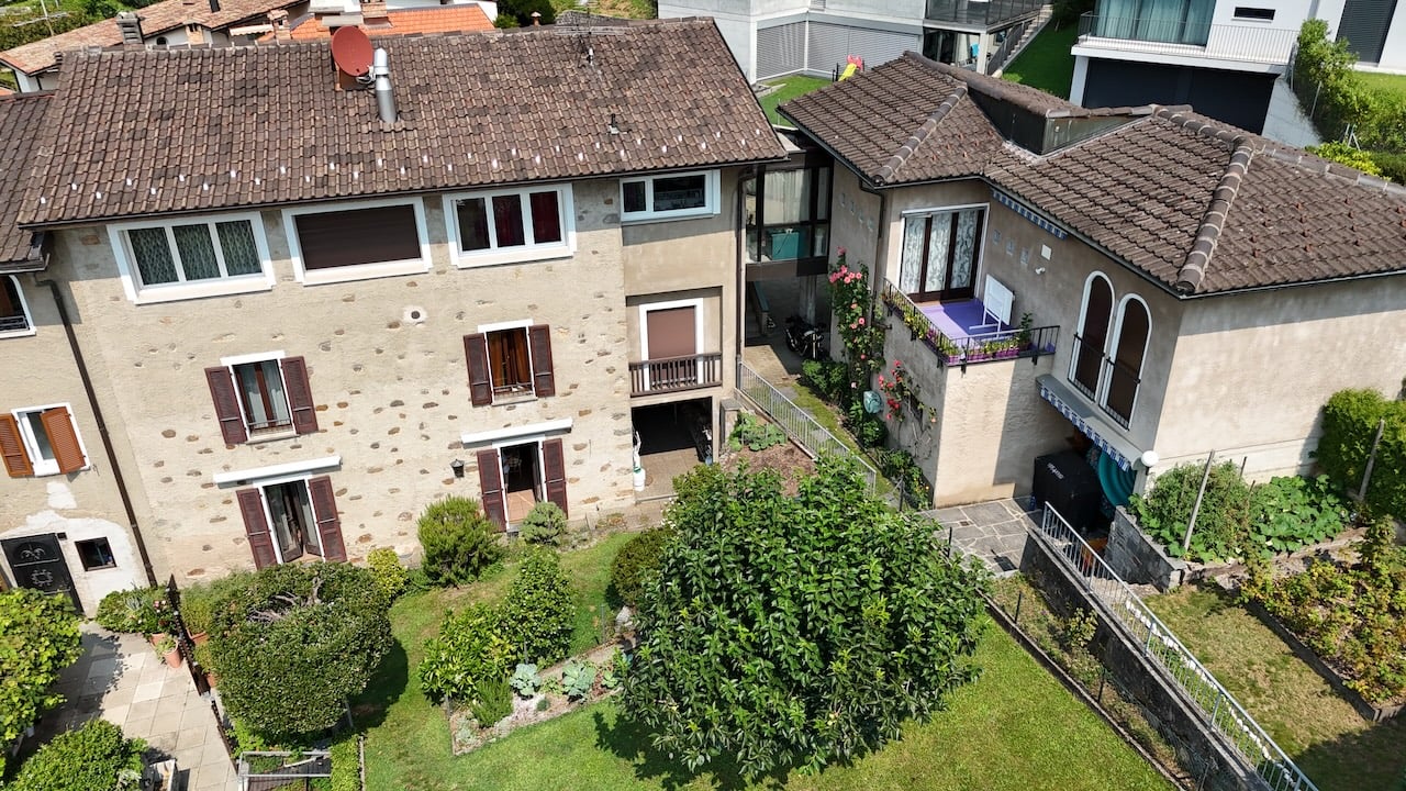 Stone exterior, tiled roof, multiple floors, several windows, balcony on the second floor, surrounded by plants and trees, paved walkway, satellite dish on the roof, nearby houses, grassy area, stairs leading to the upper floors, garden area with flowers