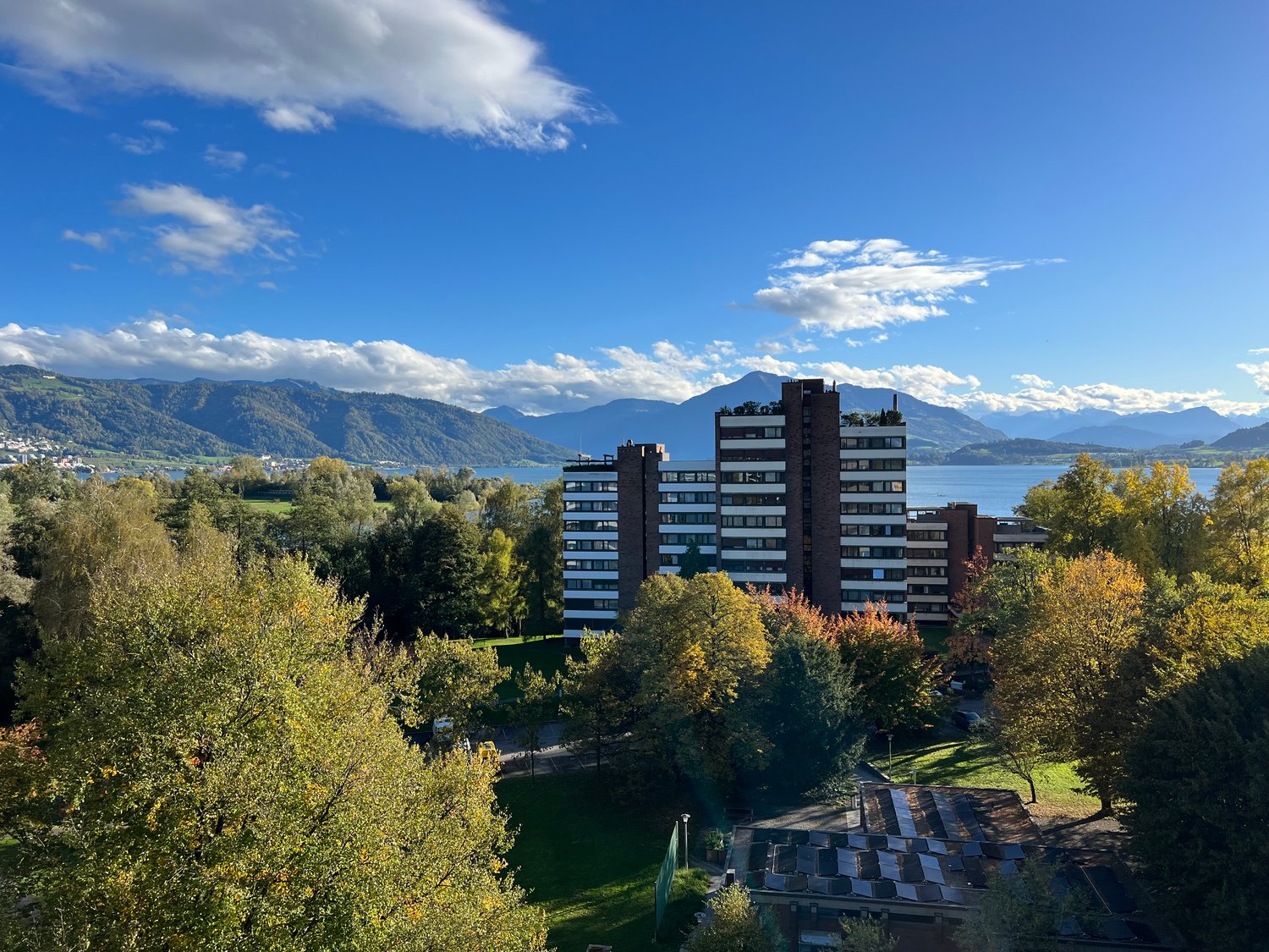 High-rise residential building with many windows, surrounded by trees and greenery, overlooking mountains and a lake.