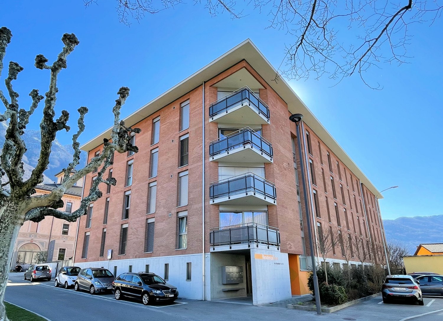 4-story brick apartment building with balconies, located on a street with parked cars